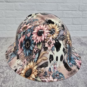 May include: A hard hat with a floral and cowhide pattern. The hat features sunflowers, pink and blue flowers, and a cowhide print. The brim is wide and the overall color scheme is neutral with pops of color.