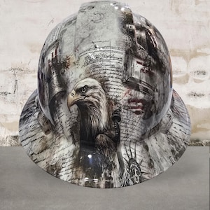 May include: A hard hat with a patriotic design featuring an eagle, the Statue of Liberty, and historical documents. The color scheme is primarily gray, white, and black, with subtle red accents. The hat has a glossy finish.