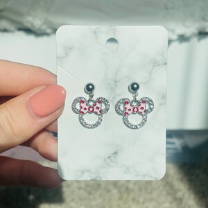 Pu&ograve; includere: Orecchini a bottone argentati con design di Minnie Mouse. Ogni orecchino presenta un fiocco rosso e bianco a pois e un contorno di cristalli trasparenti. Gli orecchini sono esposti su una carta bianca con motivo marmoreo.