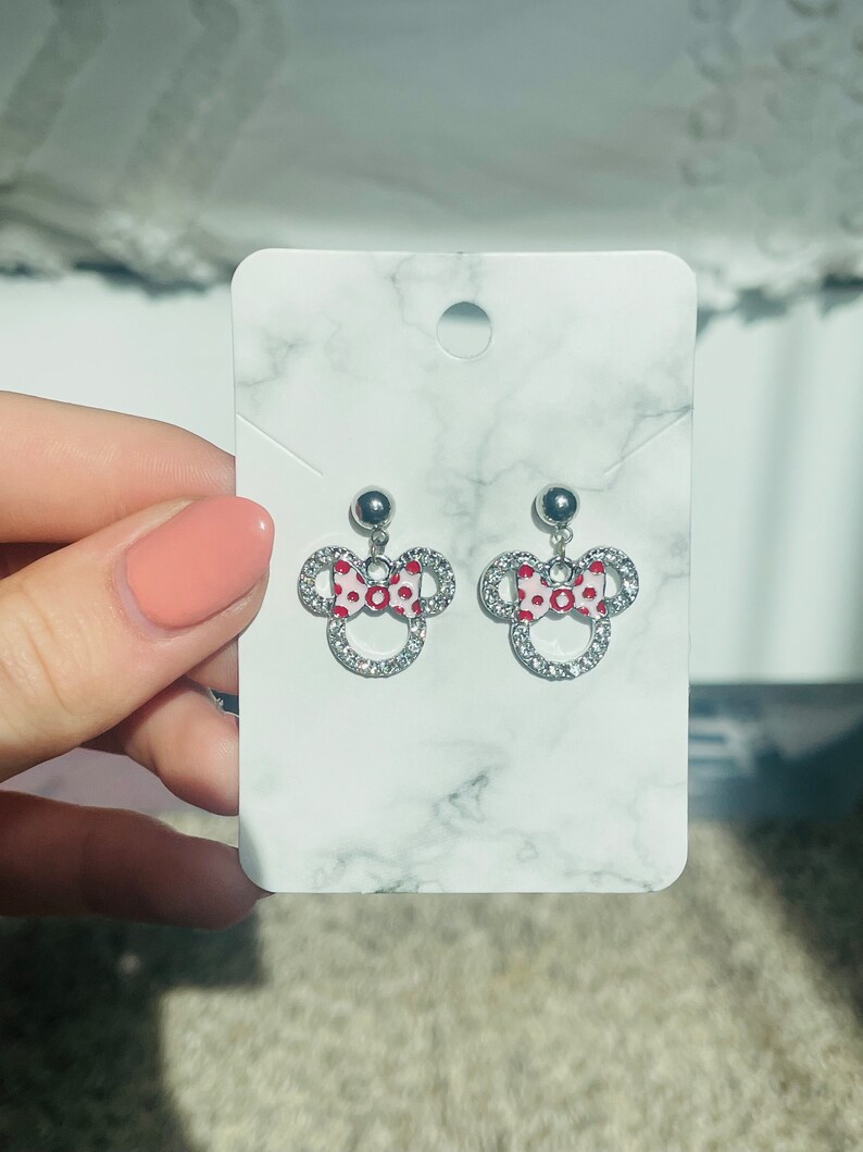 Pu&ograve; includere: Orecchini a bottone argentati con design di Minnie Mouse. Ogni orecchino presenta un fiocco rosso e bianco a pois e un contorno di cristalli trasparenti. Gli orecchini sono esposti su una carta bianca con motivo marmoreo.