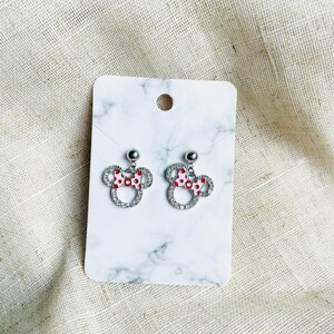 Pu&ograve; includere: Un paio di orecchini a bottone argentati a forma di testa di topo con un fiocco a pois rossi e bianchi. Gli orecchini sono incastonati con cristalli trasparenti e sono esposti su una carta bianca con motivo marmorizzato.