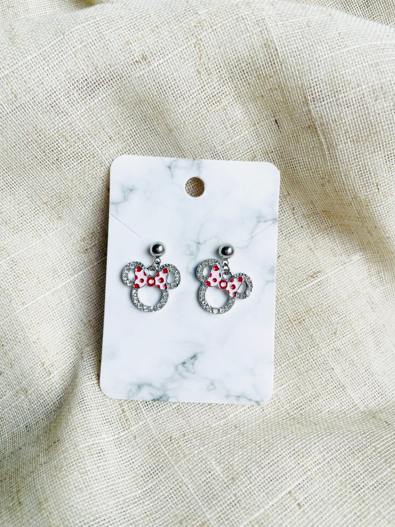 Pu&ograve; includere: Un paio di orecchini a bottone argentati a forma di testa di topo con un fiocco a pois rossi e bianchi. Gli orecchini sono incastonati con cristalli trasparenti e sono esposti su una carta bianca con motivo marmorizzato.