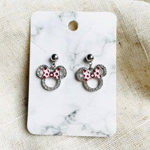 Pu&ograve; includere: Un paio di orecchini a bottone argentati a forma di testa di topo con un fiocco a pois rossi e bianchi. Gli orecchini sono impreziositi da strass trasparenti e sono esposti su una carta bianca con motivo marmorizzato.