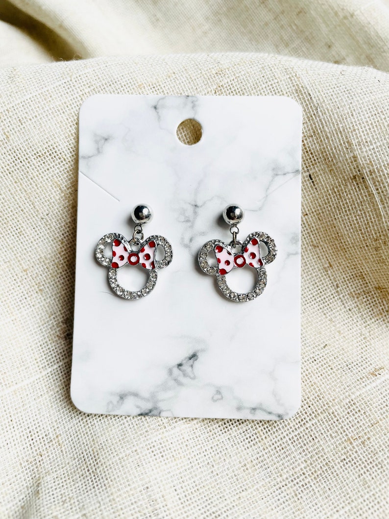 Pu&ograve; includere: Un paio di orecchini a bottone argentati a forma di testa di topo con un fiocco a pois rossi e bianchi. Gli orecchini sono impreziositi da strass trasparenti e sono esposti su una carta bianca con motivo marmorizzato.