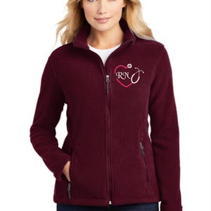 Peut inclure: Veste en polaire bordeaux avec fermeture éclair et deux poches zippées. La veste est ornée d'un cœur brodé blanc avec les lettres "RN" et un stéthoscope à l'intérieur du cœur.