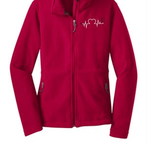 Peut inclure: Une veste en polaire rouge avec une fermeture éclair et deux poches. La veste a un graphique de battement de cœur blanc avec un stéthoscope sur la poitrine gauche.