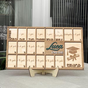 Puede incluir: Tablero de exhibición de años escolares de madera con ranuras para fotos. Presenta el nombre "Luca Cruise", un birrete y la palabra "Senior". Incluye el texto "Hello World", "1 año" a "11º grado".