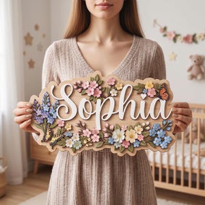 Op de afbeelding: Een houten wandbord met de naam Sophia in witte schrijfletters. Het bord is versierd met gelaagde, kleurrijke bloemstukken, waaronder roze, blauwe en gele bloemen, samen met twee vlinders, één oranje en één geel van kleur.
