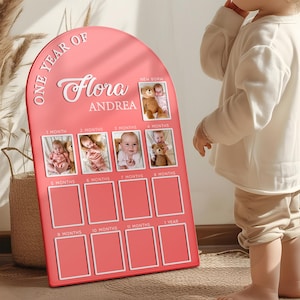 Peut inclure: Un tableau d'affichage photo en forme d'arche, de couleur corail, avec le texte "ONE YEAR OF" et le nom "Flora Andrea". Il comporte des espaces pour les photos de la naissance à un an. Le tableau comprend les mois et un espace pour une photo de nouveau-né.