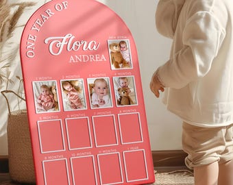 May include: A coral-colored, arched photo display board with the text "ONE YEAR OF" and the name "Flora Andrea." It has spaces for photos from newborn to one year. The board includes the months and a space for a newborn photo.