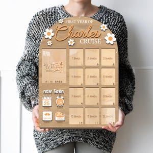Puede incluir: Tablero de hitos de madera con el texto "First Year of Charles Cruise" y detalles de margaritas. Incluye espacios para fotos e hitos mensuales. Presenta el texto "Welcome to the World" y los datos de nacimiento del bebé: fecha, hora, peso y altura (53 cm).