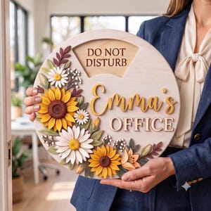 May include: A round wooden sign featuring the text DO NOT DISTURB, Emma's, and OFFICE in raised lettering. The sign is decorated with layered, three-dimensional floral elements including yellow sunflowers, white daisies, and green leaves.