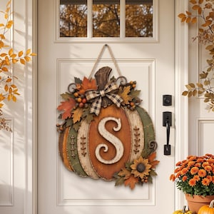 May include: A decorative pumpkin-shaped door hanger featuring a large white letter S in the center. The pumpkin is painted in shades of orange and green with stitched detailing, adorned with a black and white plaid bow, autumn leaves, and berries.