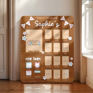 Może przedstawiać: Drewniana tablica milowa dla dziecka z napisem "First Year of Sophie's". Posiada miejsca na zdjęcia, grafikę "Welcome to the World" oraz sekcje na dane urodzenia dziecka i miesięczne kamienie milowe, ozdobiona kwiatami i sercami.