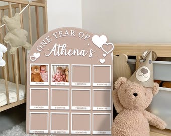 May include: A taupe-colored baby milestone photo board with the text "ONE YEAR OF Athena's". The board has spaces for 12 photos, with the months labeled. Two photos are visible. Decorative hearts are at the top.