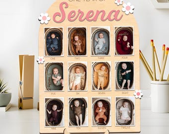 May include: A wooden photo display for a baby's first year, featuring twelve small photos. The display is arched at the top with the name "Serena" and the phrase "ONE YEAR OF" in pink script. White flower accents are placed around the edges.