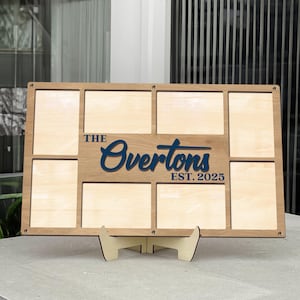 Puede incluir: Un expositor de fotos familiar de madera con ocho aberturas rectangulares. La pantalla presenta el texto "The Overtons EST. 2025" en azul marino. Se apoya en un soporte de madera y está diseñado para contener varias fotos.