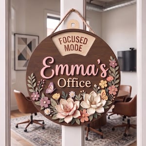Puede incluir: Un letrero redondo de madera con acabado marrón oscuro, que presenta el texto "FOCUSED MODE" en la parte superior y "Emma's Office" en el centro. El letrero está decorado con flores, hojas y dos mariposas en tonos pastel de rosa, crema y verde.