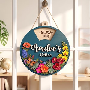 May include: A circular dark teal wooden sign hanging by a rope, featuring the text "FORCUSED MODE" and "Amelia's Office" in white lettering. The sign is decorated with colorful 3D floral elements, including red, yellow, and pink flowers, green succulents, and a monarch butterfly.