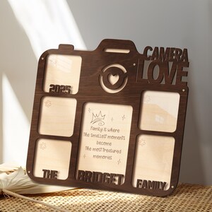 May include: A wooden photo frame in the shape of a camera, with spaces for multiple photos. The frame is dark brown with the words "CAMERA LOVE" cut out at the top. The text "2025", "THE", "BRIDGET", and "FAMILY" are also visible. The frame includes the quote: "Family is where the smallest moments become the most treasured memories."