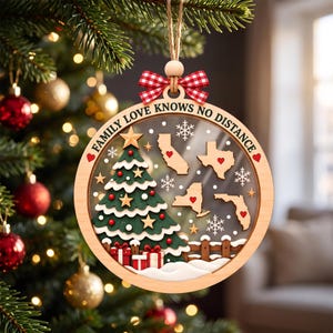 May include: A round wooden Christmas ornament featuring a green tree, red gifts, and four US state maps with red hearts. The text reads FAMILY LOVE KNOWS NO DISTANCE. It is decorated with white snowflakes, gold stars, and a red and white gingham bow.