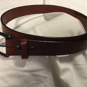 May include: A brown leather belt with a black metal buckle and two black rivets. The belt is folded in half and lying on a white surface.