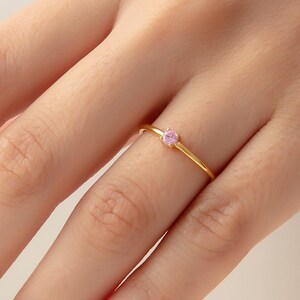 May include: A slender gold ring with a small, round pink gemstone. The ring is set on a thin, polished gold band. The ring is displayed on a finger, showcasing its simple elegance and design.