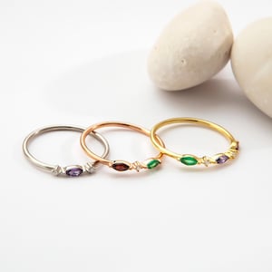 Op de afbeelding: Drie fijne ringen in zilver, roségoud en goud. Elke ring heeft een centrale edelsteen: paars, rood en groen. De ringen liggen op een witte achtergrond met twee gladde, lichte stenen.
