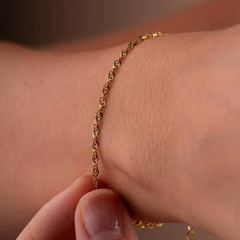Op de afbeelding: Een delicate gouden kettingarmband met een delicaat, ingewikkeld ontwerp. De armband wordt getoond op de pols van een persoon.