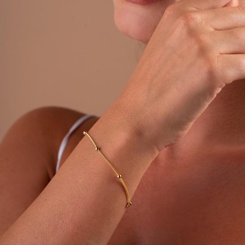 Op de afbeelding: Een delicate gouden kettingarmband met kleine ronde kralen. De armband wordt getoond op de pols van een persoon.