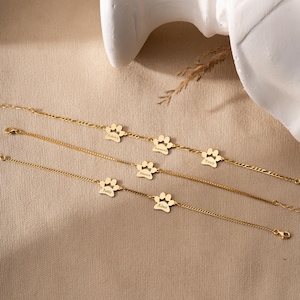 Könnte beinhalten: Fünf goldene Armbänder mit Pfotenabdruck-Anhängern, auf denen verschiedene Namen graviert sind. Die Namen sind "Abbie", "Amara", "Tony", "Damon", "Teddy" und "Alex".