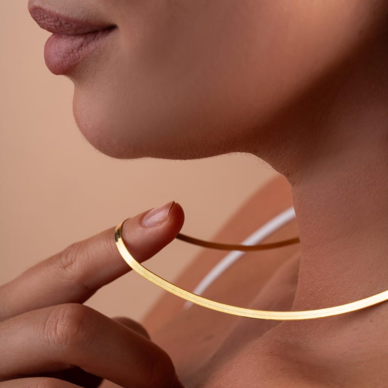 May include: A gold chain necklace with a flat, wide design. The necklace is worn by a person with their finger touching the chain.