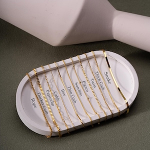 Op de afbeelding: Een close-up van een witte display met 12 verschillende gouden kettingstijlen. De kettingen zijn gelabeld met hun namen: Rope, Thicker Curb, Paperclip, Cable, Box, Thin Curb, Satellite, Figaro, Twist, Thick Curb en Snake.