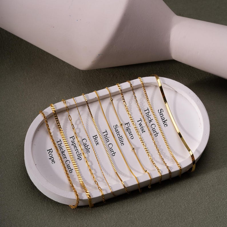 Op de afbeelding: Een close-up van een witte display met 12 verschillende gouden kettingstijlen. De kettingen zijn gelabeld met hun namen: Rope, Thicker Curb, Paperclip, Cable, Box, Thin Curb, Satellite, Figaro, Twist, Thick Curb en Snake.
