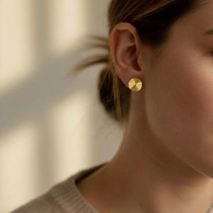 May include: A close-up of a gold-colored stud earring with a radial design. The earring is circular and has a textured surface. The earring is worn on the ear of a person wearing a light-colored sweater.