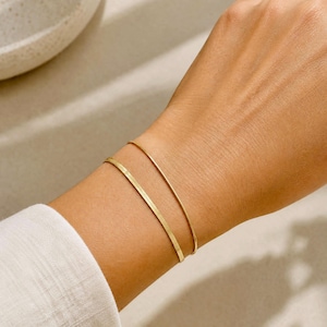 Può includere: Due braccialetti sottili color oro indossati al polso. Un braccialetto presenta un design a catena a serpente piatta e flessibile con un motivo a spina di pesce visibile, mentre il secondo è un semplice bracciale a filo color oro, liscio e sottile.