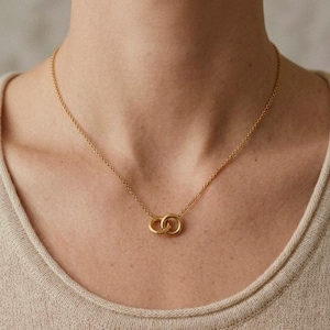 May include: A gold necklace featuring a pendant of two intertwined circles. The chain is delicate, and the pendant sits at the neckline. The background is a plain, neutral colour.