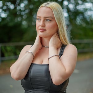 Może przedstawiać: Srebrny naszyjnik typu choker i pasujące srebrne bransoletki. Biżuteria jest noszona z czarnym topem ze skóry. Kobieta na zdjęciu ma blond włosy i jest na zewnątrz.