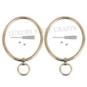 Op de afbeelding: Twee goudkleurige metalen ringen met een kleinere ring aan de onderkant. Elke ring heeft een klein zilveren gereedschap en twee kleine zilveren pinnen. De tekst "LUXURY STEEL CRAFTS" staat op de ringen.