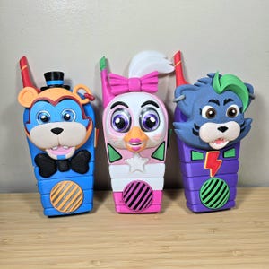 May include: Three colorful toy walkie-talkies featuring cartoon animal faces. The walkie-talkies are blue, pink, and purple, with orange, green, and white accents. Each has a unique animal design, including a bear, a chicken, and a wolf.