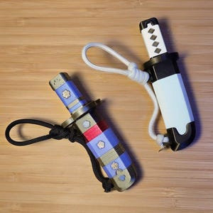 May include: Two miniature katana keychains. One is blue, red, and gold with a black cord. The other is white and black with a white cord. Both have decorative floral accents and are set on a wooden surface.