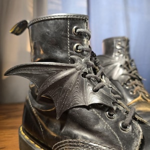 May include: Black leather boots with bat wing accessories. The bat wings are a dark gray color and are attached to the laces of the boots. The boots have a classic design with visible stitching and metal eyelets.