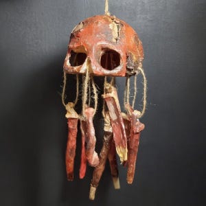 May include: A hanging mobile featuring a weathered, reddish-brown skull at the top. Several long, thin, textured, and reddish-brown bone-like appendages are suspended from the skull by natural-colored twine, creating a layered, dangling structure.
