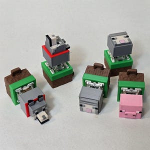 May include: A collection of Minecraft-themed figurines. The figures include a wolf, pig, and sheep, each with a blocky design. The figurines are gray, pink, and white, with brown and green accents. The figures are on green and brown bases.