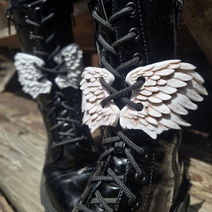 May include: Black, glossy boots with white, textured wing-shaped embellishments laced through the front. The wings are detailed with feather-like textures and are attached to the laces. The boots have black laces and a platform sole.