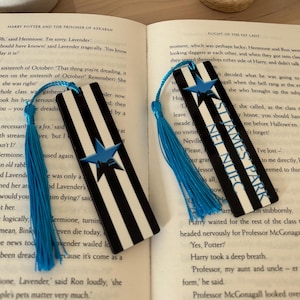 K&ouml;nnte beinhalten: Zwei schwarz-wei&szlig; gestreifte Lesezeichen mit blauen Quasten und einem blauen Sterndesign werden auf einem offenen Buch gezeigt. Die Lesezeichen tragen die Texte "NEL NUFC" und "ST JAMES PARK". Eine Kerze, eine kleine Sukkulente und ein Schild mit dem Text "RMR DESIGNS" befinden sich im Hintergrund.