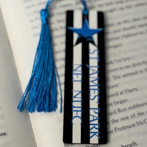 K&ouml;nnte beinhalten: Lesezeichen mit blauer Quaste, schwarzen und wei&szlig;en Streifen und einem blauen Stern. Der Text "ST JAMES PARK NUFC" ist vertikal aufgedruckt. Das Lesezeichen liegt auf einem offenen Buch.