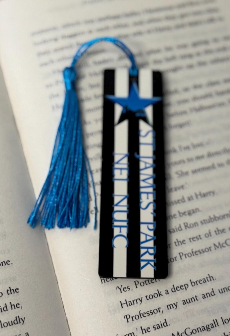 K&ouml;nnte beinhalten: Lesezeichen mit blauer Quaste, schwarzen und wei&szlig;en Streifen und einem blauen Stern. Der Text "ST JAMES PARK NUFC" ist vertikal aufgedruckt. Das Lesezeichen liegt auf einem offenen Buch.