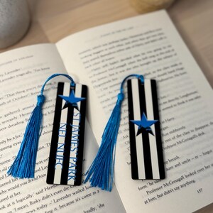 K&ouml;nnte beinhalten: Zwei rechteckige Lesezeichen mit schwarz-wei&szlig;en Streifen, einem blauen Stern und dem Text "ST JAMES PARK". Jedes Lesezeichen hat eine blaue Quaste. Die Lesezeichen liegen auf einem offenen Buch.
