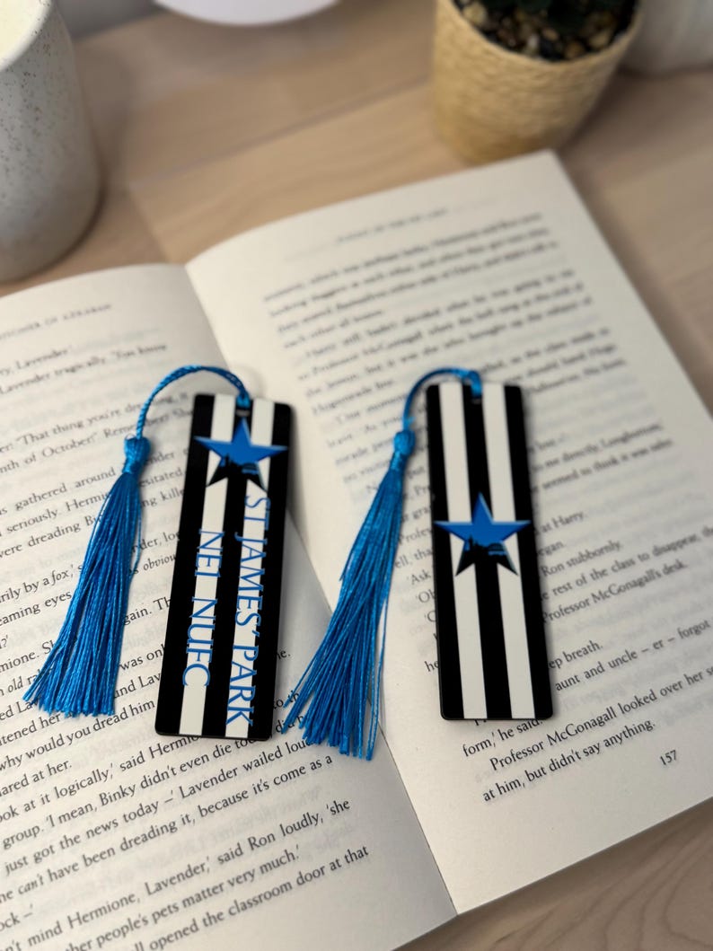 K&ouml;nnte beinhalten: Zwei rechteckige Lesezeichen mit schwarz-wei&szlig;en Streifen, einem blauen Stern und dem Text "ST JAMES PARK". Jedes Lesezeichen hat eine blaue Quaste. Die Lesezeichen liegen auf einem offenen Buch.